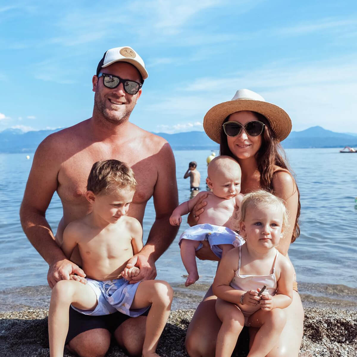 A family of five poses at the beach with mountains in the background; two adults and three young children.