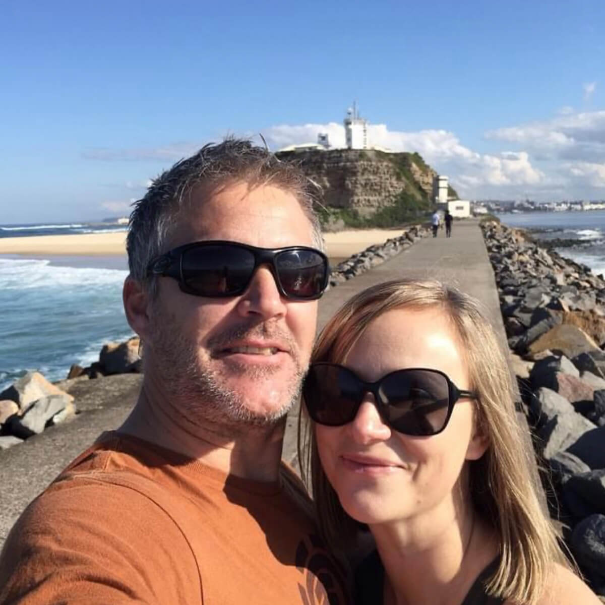 A couple poses for a selfie on a rocky path by the ocean, with a hill and lighthouse in the background.