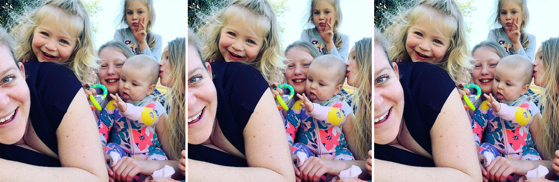 A group of smiling children and a woman pose together outdoors, with one baby playing with a toy.
