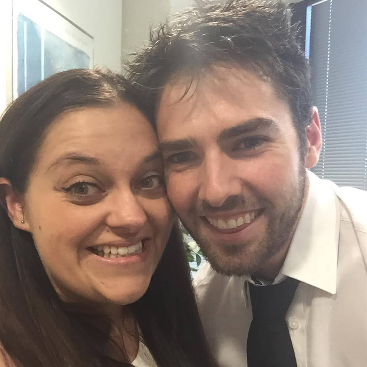 A smiling woman and man pose closely together for a selfie, with indoor plants and window blinds in the background.