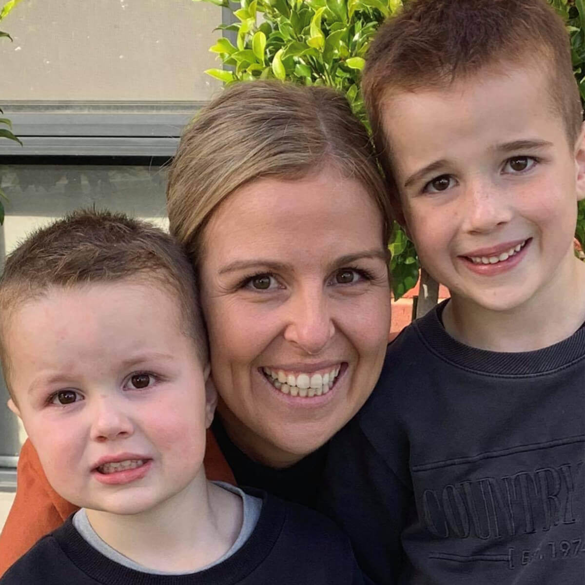 A woman smiles with two young boys, one on each side, against a backdrop of green foliage.