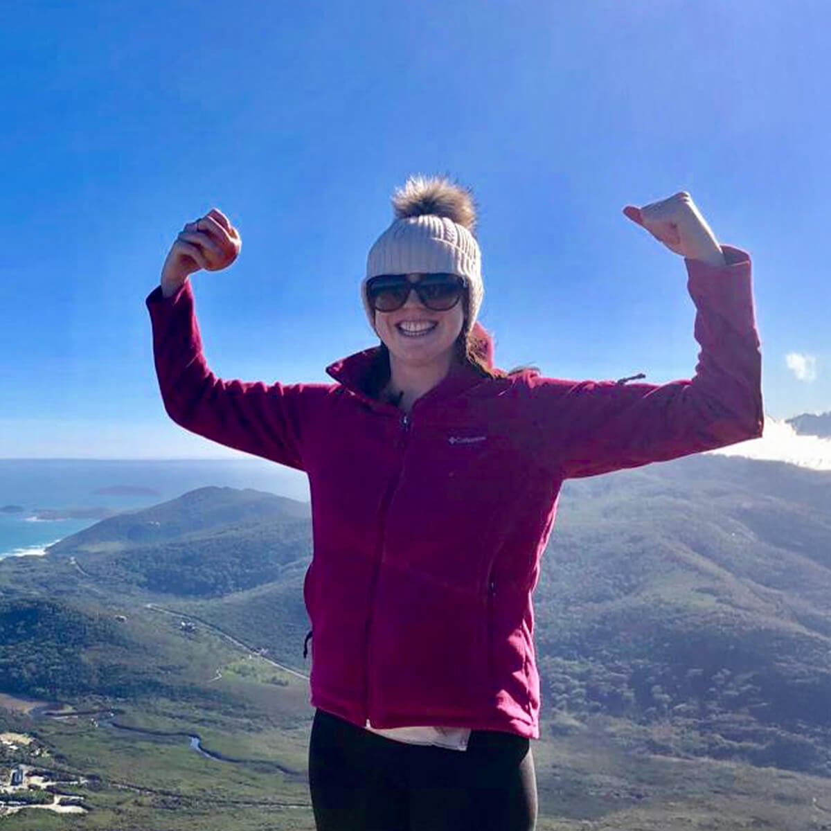 A smiling woman in a pink jacket and sunglasses raises her arms triumphantly against a scenic mountain and ocean background.