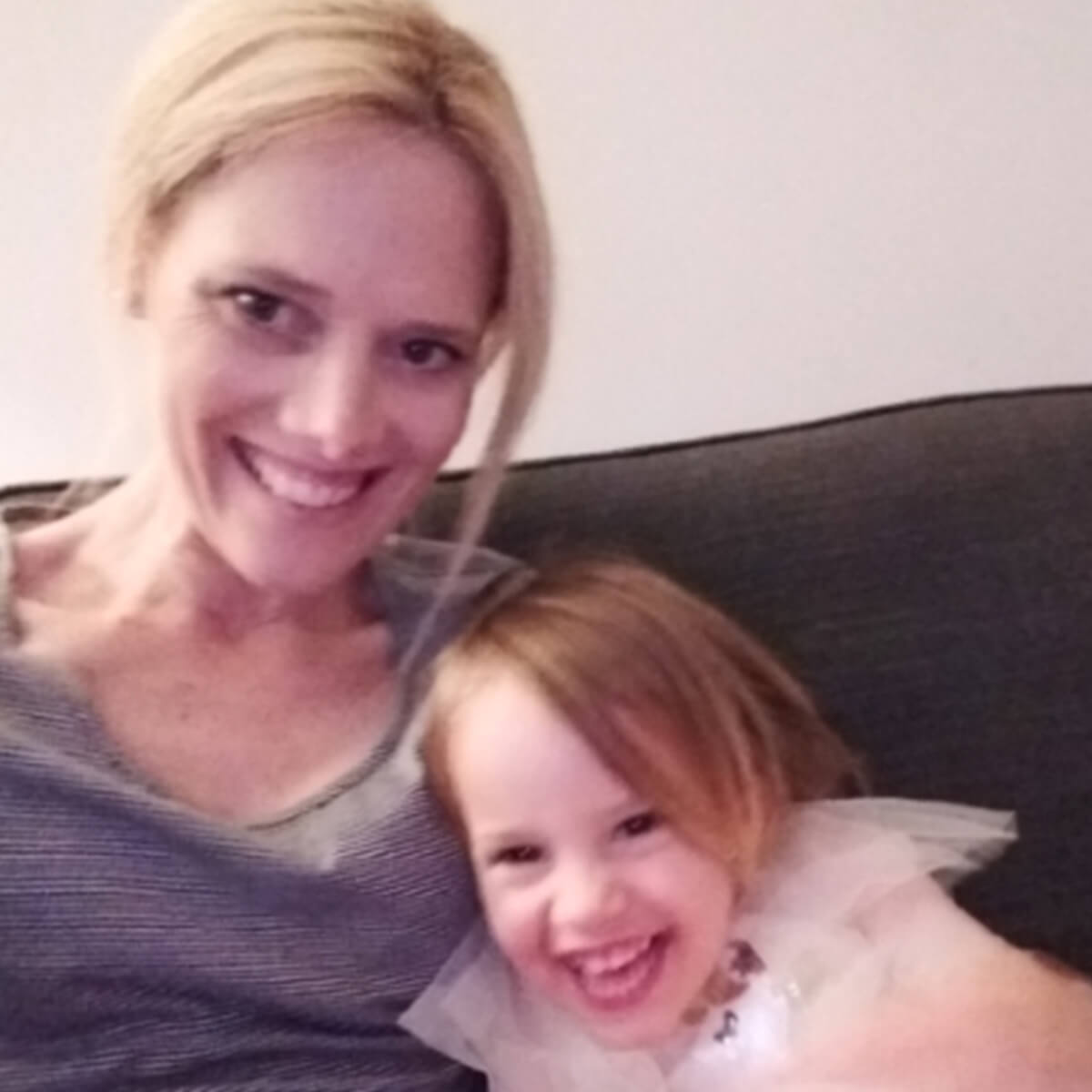 A smiling woman and a young girl are sitting together on a couch, both looking happy and playful.