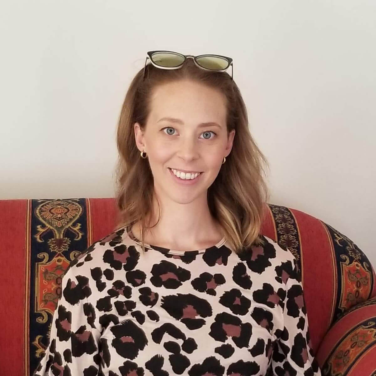 A woman with light brown hair, wearing a leopard print top, smiles while sitting on a patterned couch.