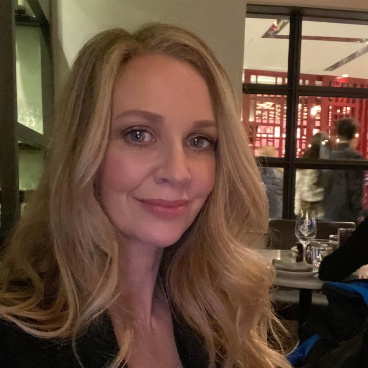 A woman with long, wavy blonde hair smiles at the camera in a restaurant setting.