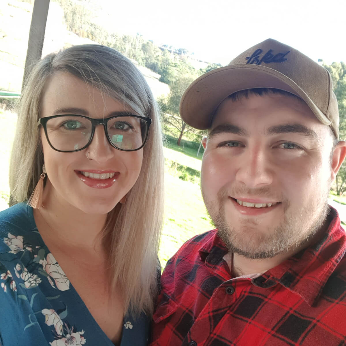 A smiling woman with glasses and a man in a plaid shirt pose together outdoors with greenery in the background.