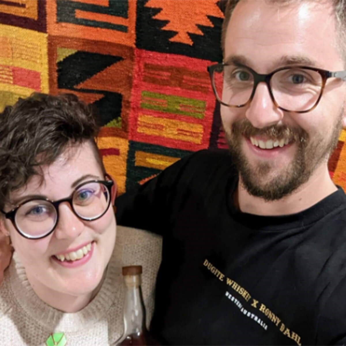 Two smiling individuals pose for a selfie in front of a colorful patterned wall, holding a bottle.