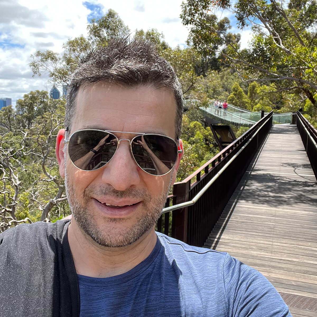 A man in sunglasses smiles for a selfie on a wooden walkway surrounded by trees, with people in the background.