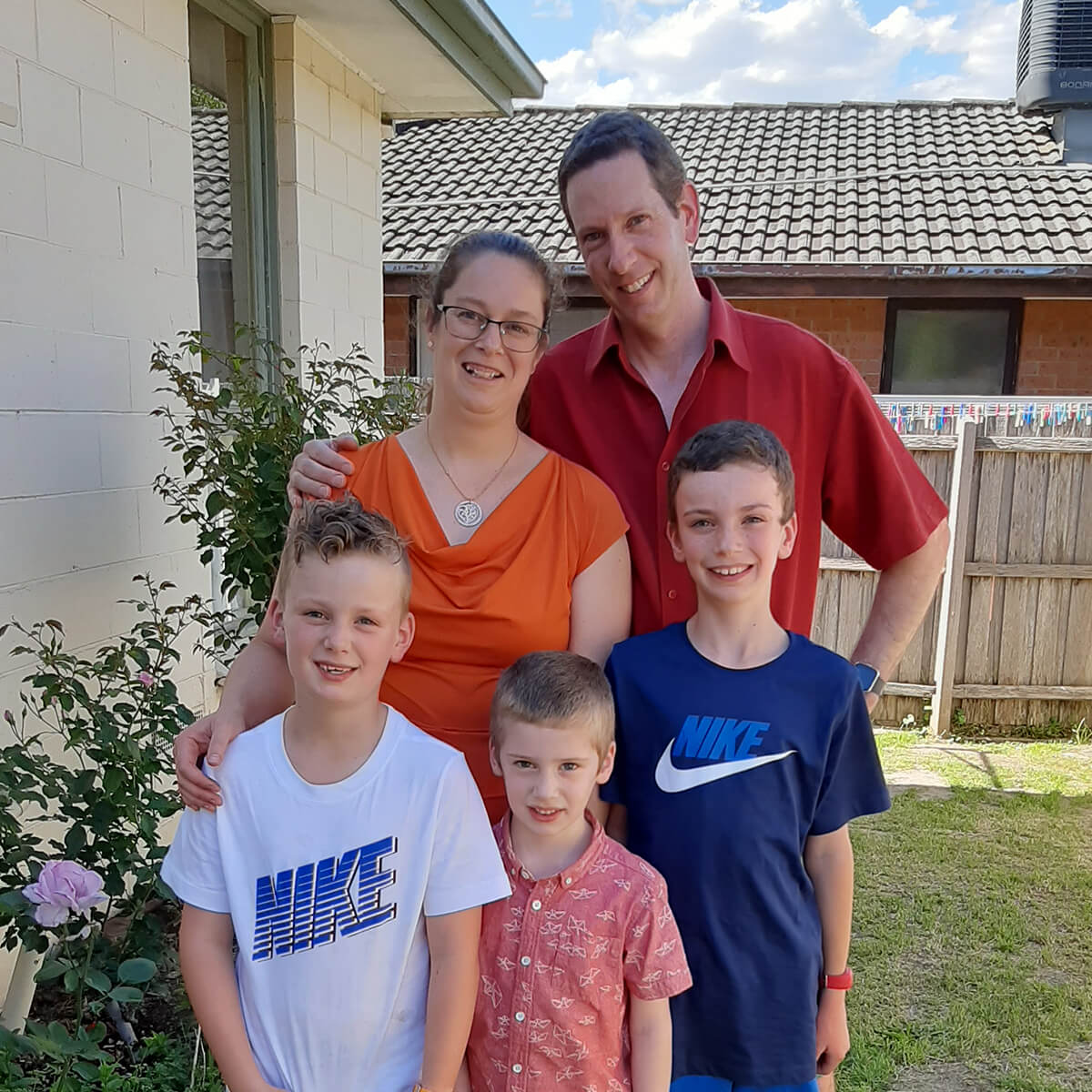 A family of five poses outdoors; two adults and three children, smiling in front of a house with greenery.