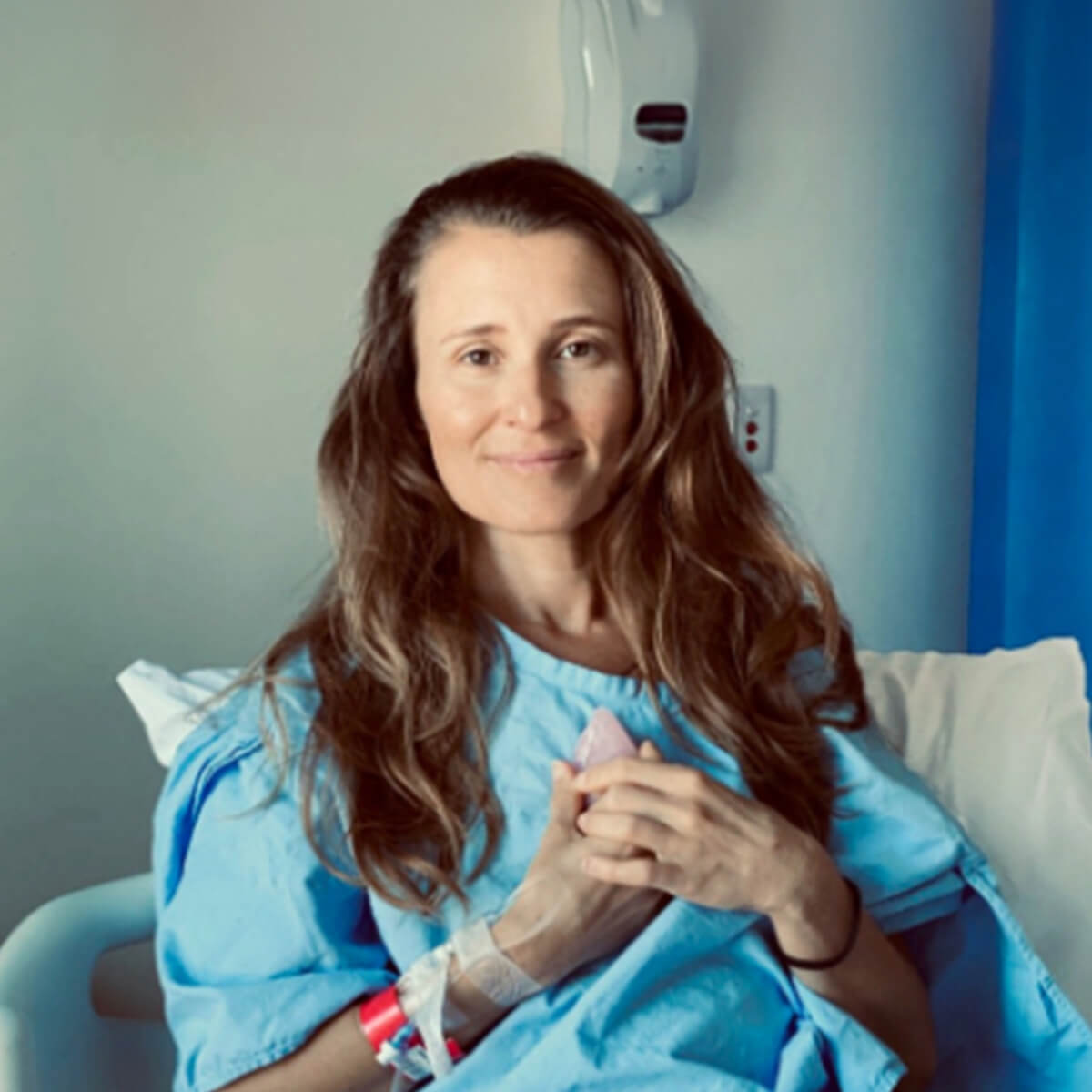 A woman in a hospital gown sits on a bed, smiling and holding a small pink object in her hands.