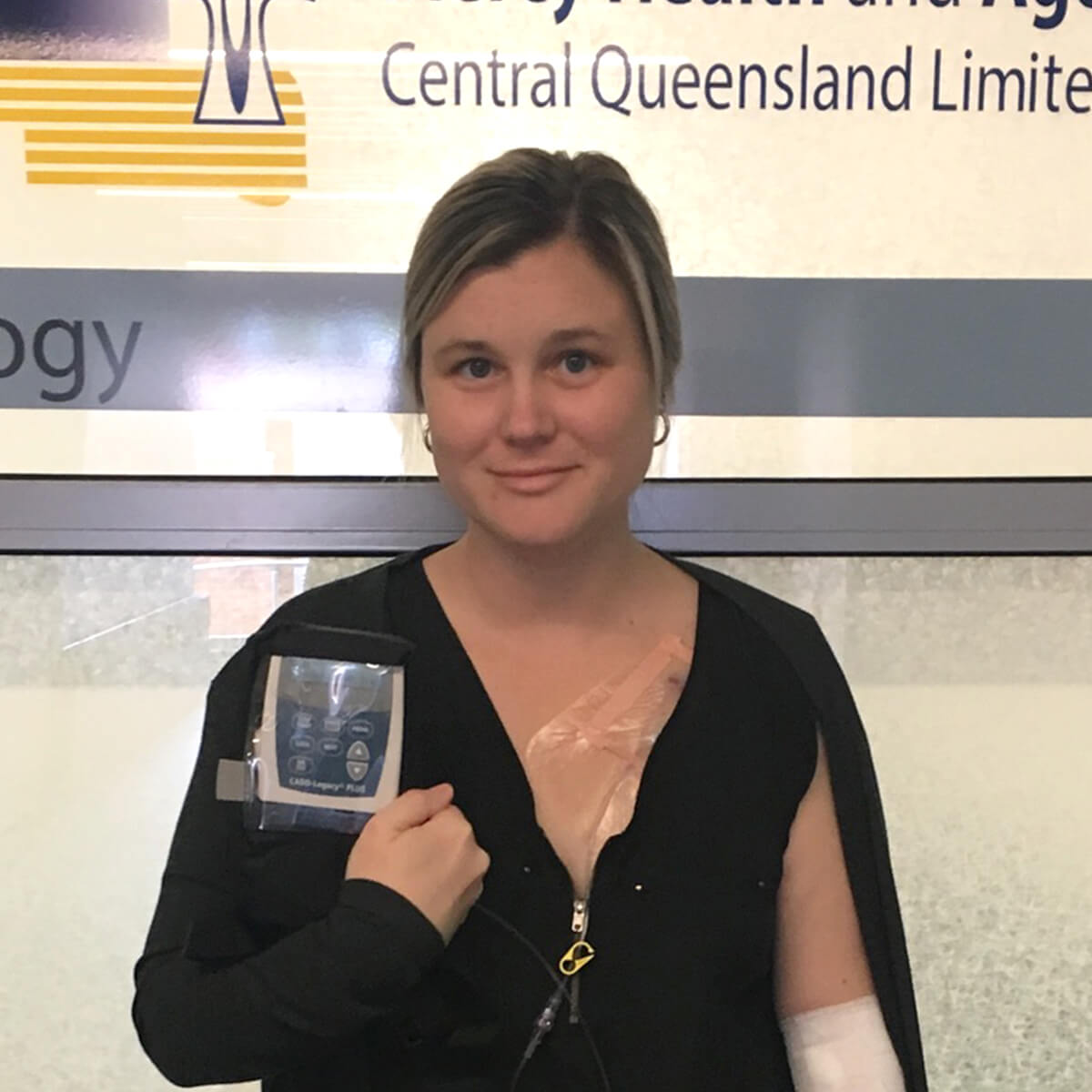 A woman in a black top stands in front of a sign, holding a medical device attached to her shoulder.