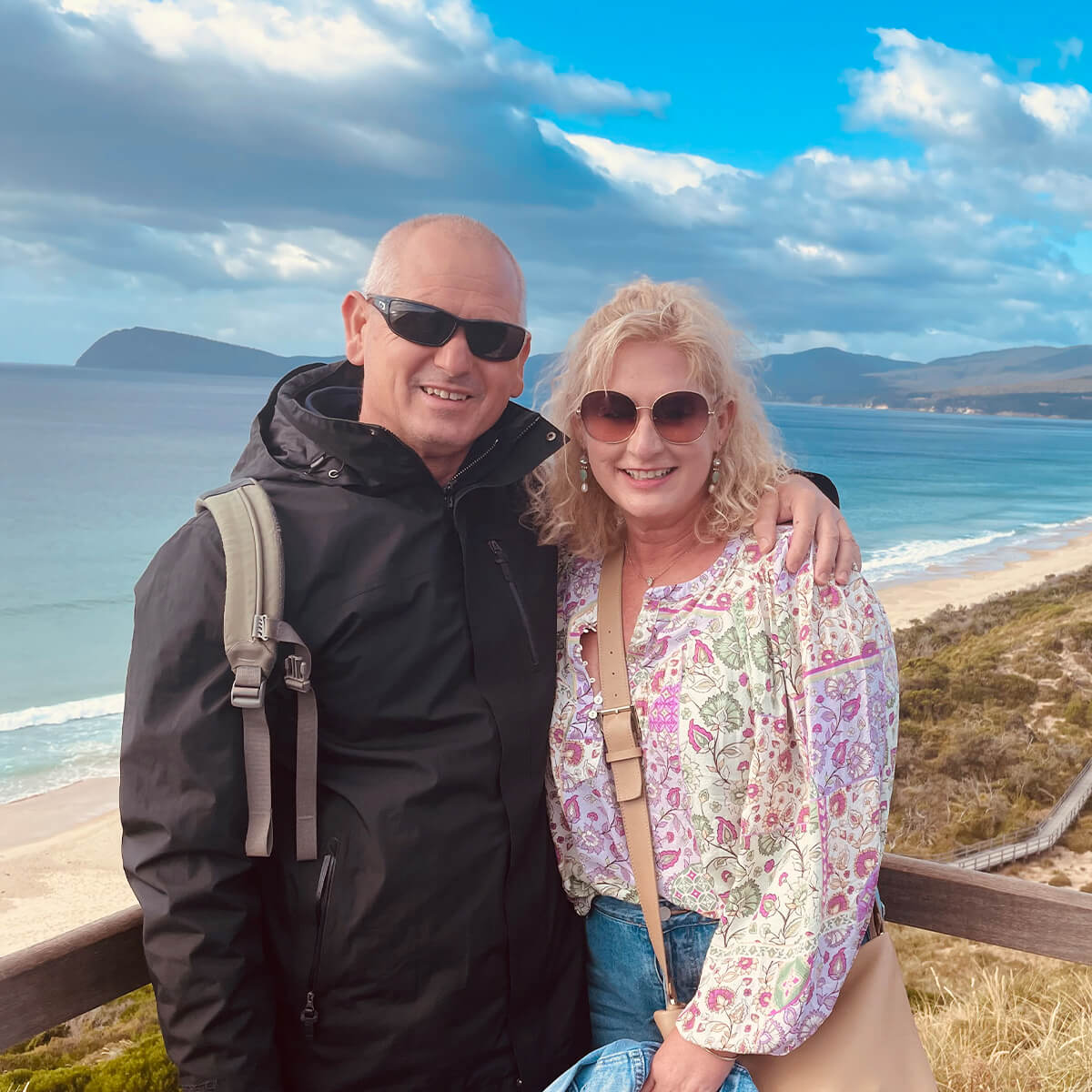 A man and woman stand together on a coastal overlook, smiling with a beach and blue ocean in the background.