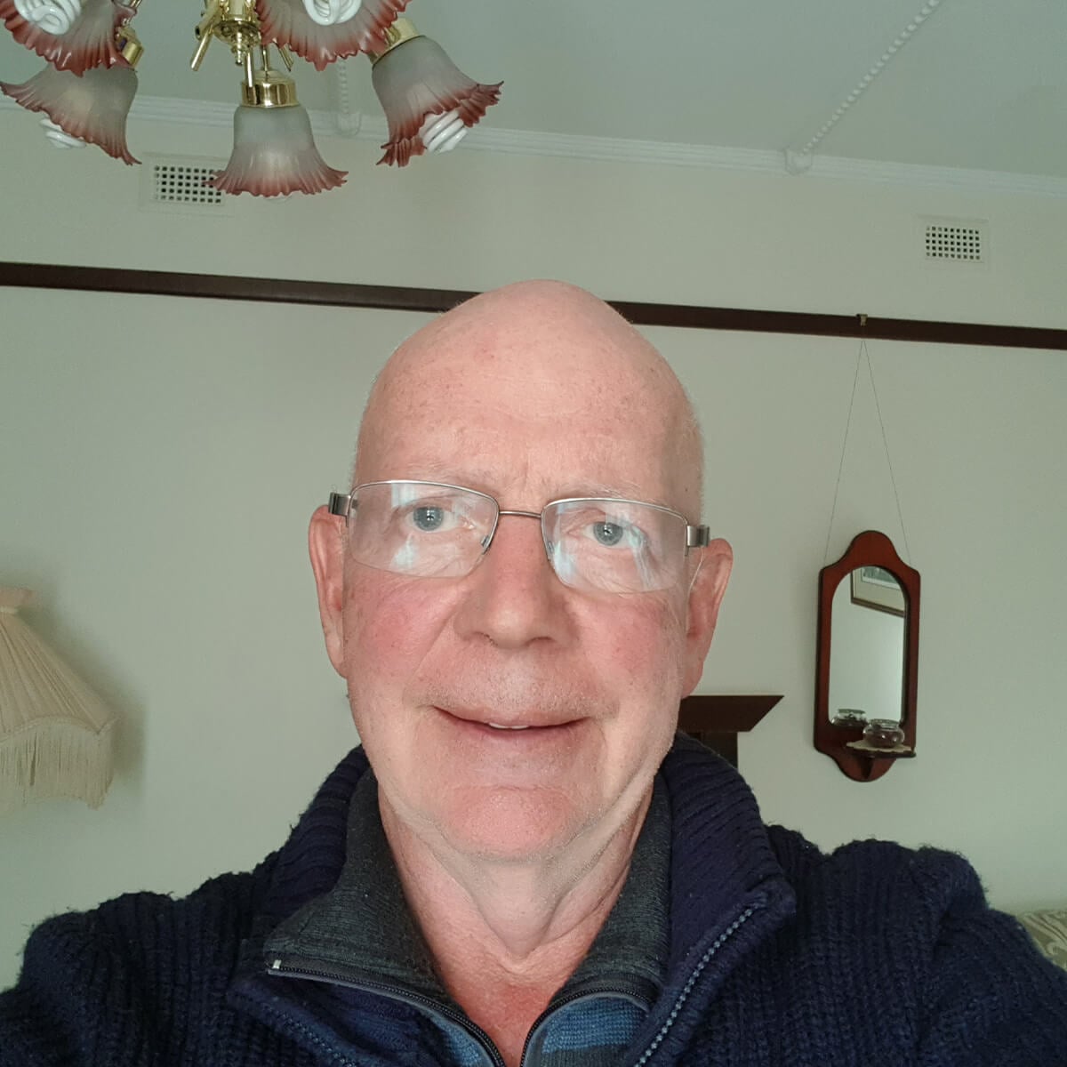 A man with glasses and a bald head smiles at the camera, with a chandelier and a small mirror visible in the background.