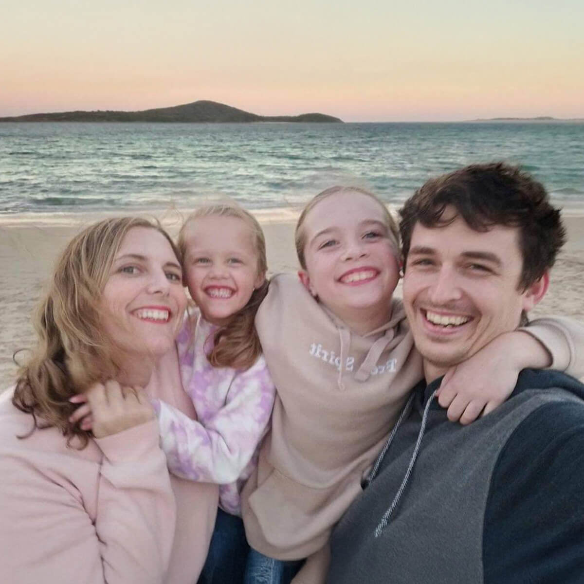 A family of four smiles for a selfie on the beach at sunset, with water and hills in the background.