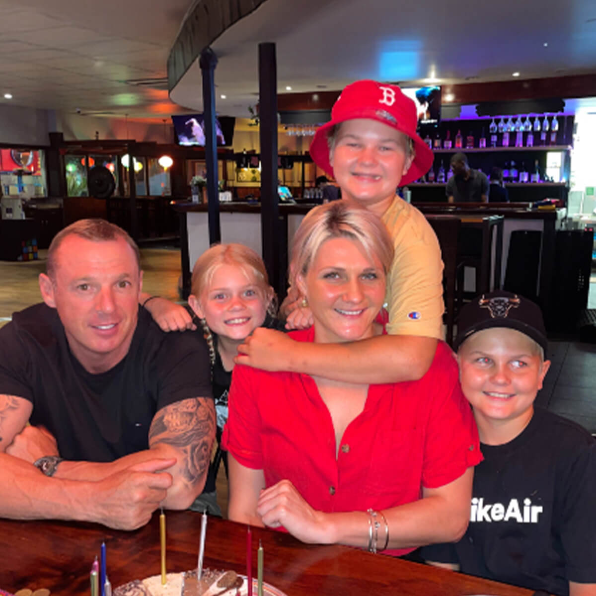A family of five poses together at a restaurant, smiling with a birthday cake and candles in front of them.