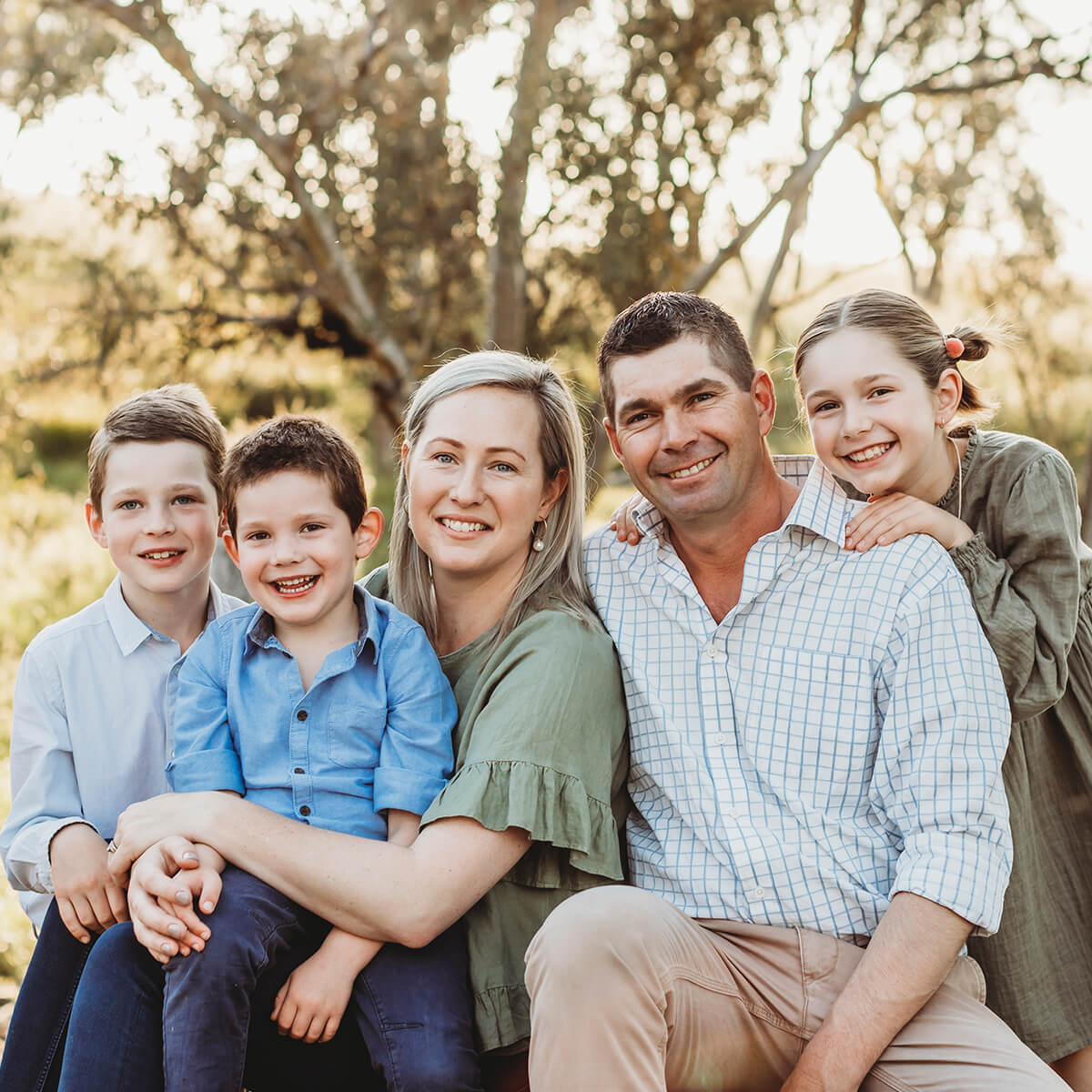A family of five smiles together outdoors, with two boys, a girl, and their parents sitting in a natural setting.