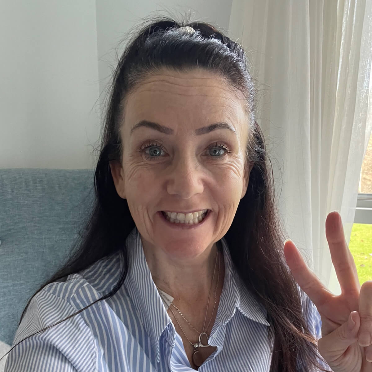 A woman with long dark hair smiles and makes a peace sign, wearing a blue and white striped shirt.