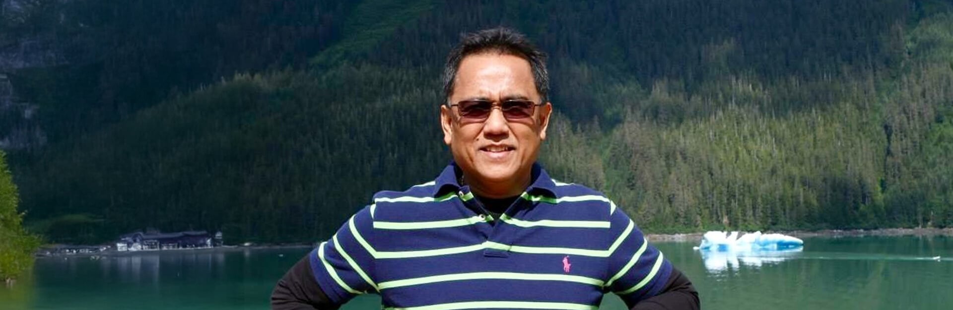 A man in a striped polo shirt stands with hands on hips, surrounded by mountains and a lake with icebergs.