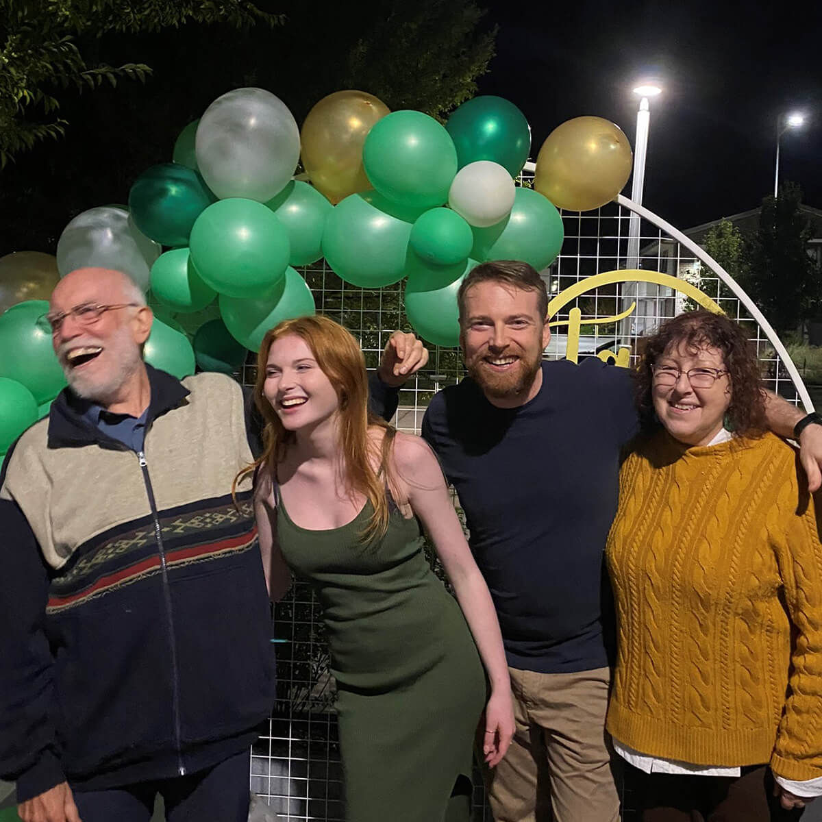 Four people smile and pose together in front of a backdrop of green and gold balloons at night.