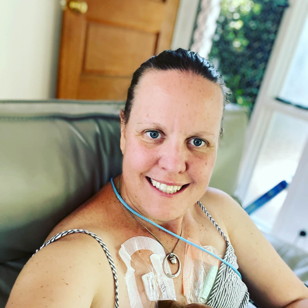 A smiling woman with short hair, wearing a striped top, sits on a couch with a medical device attached to her chest.