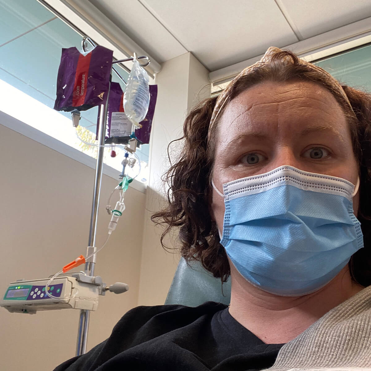 A person wearing a blue mask sits in a medical setting with an IV stand and bags in the background.