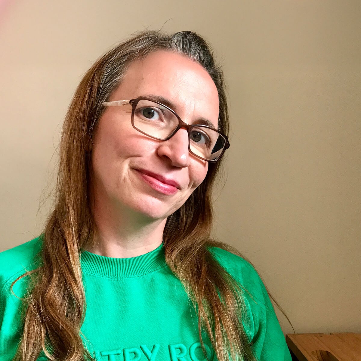 A woman with long hair and glasses smiles, wearing a green sweatshirt against a neutral background.