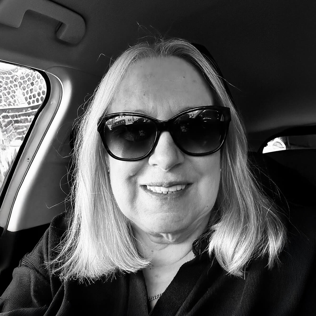 A woman with long, straight hair and sunglasses smiles from the driver's seat of a car.