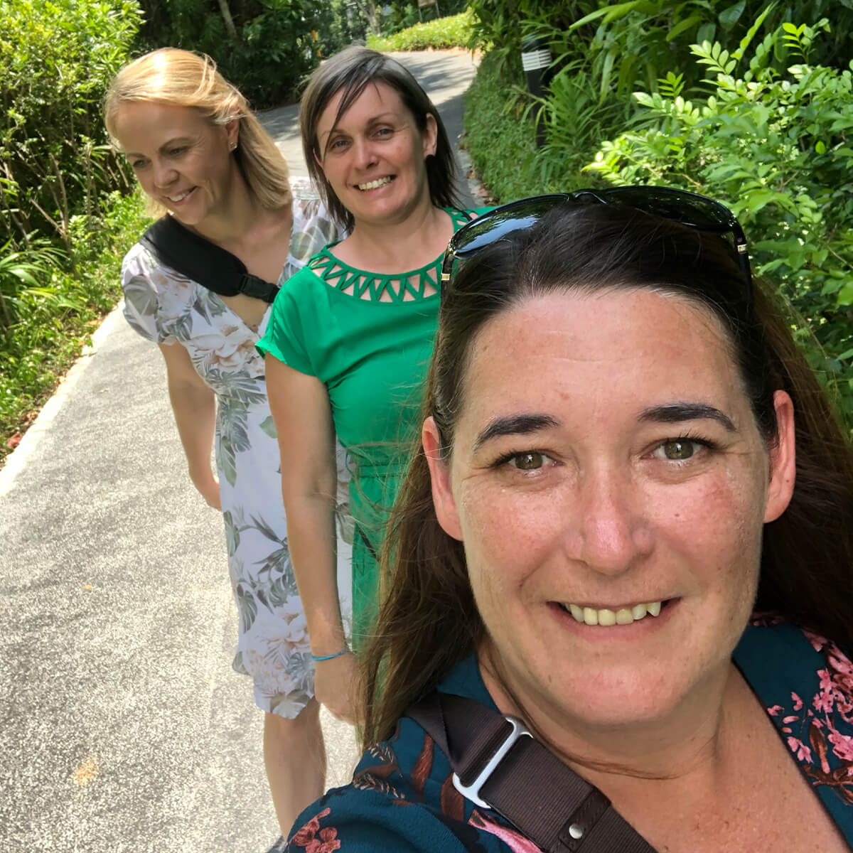 Three women smile while walking along a path surrounded by greenery. One woman takes a selfie in the foreground.