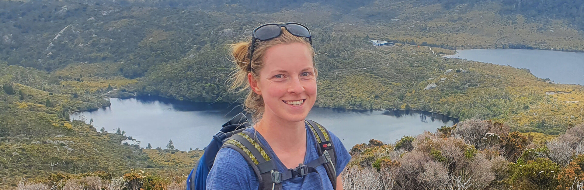 A woman with a backpack smiles in front of a scenic landscape featuring lakes and greenery.