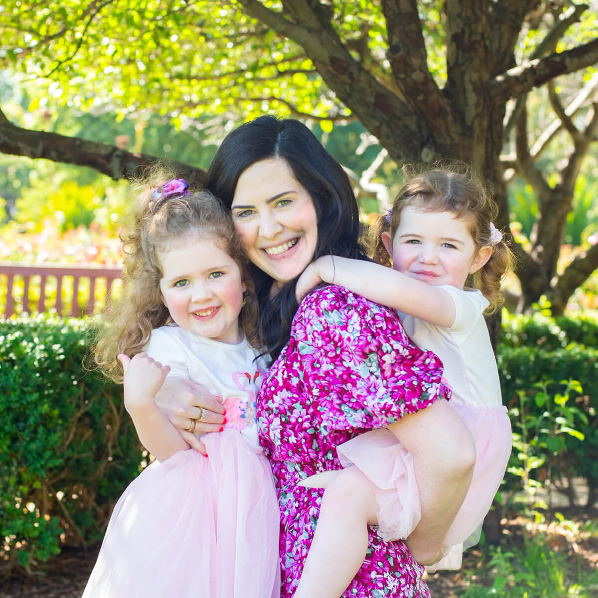 A woman smiles while holding two young girls, all dressed in light clothing, in a sunny garden setting.