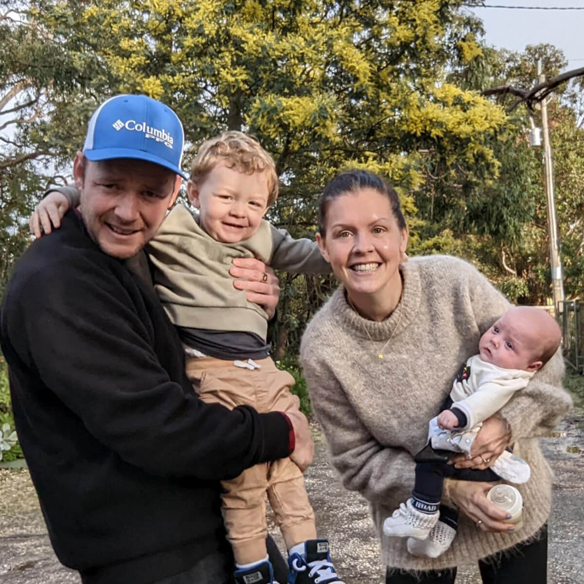 A man holds a smiling toddler, while a woman holds a baby. They are outdoors with greenery in the background.