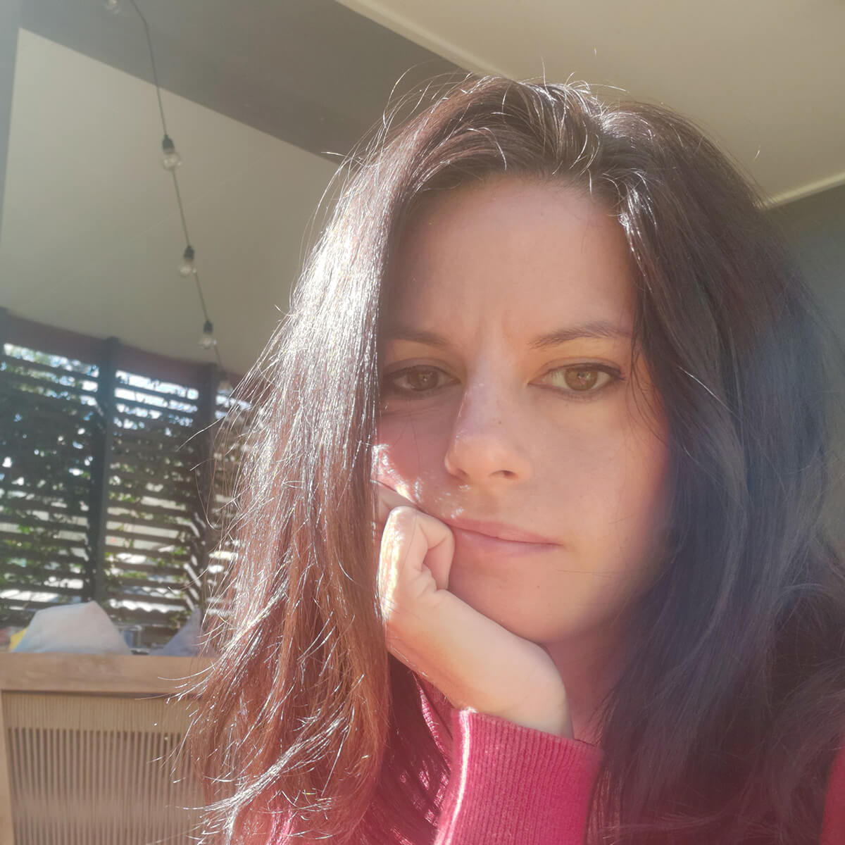A woman with long brown hair rests her chin on her hand, looking thoughtfully at the camera in a sunlit setting.
