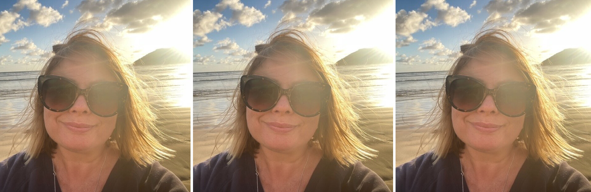 A person with sunglasses smiles at the beach, with sunlight and clouds in the background.