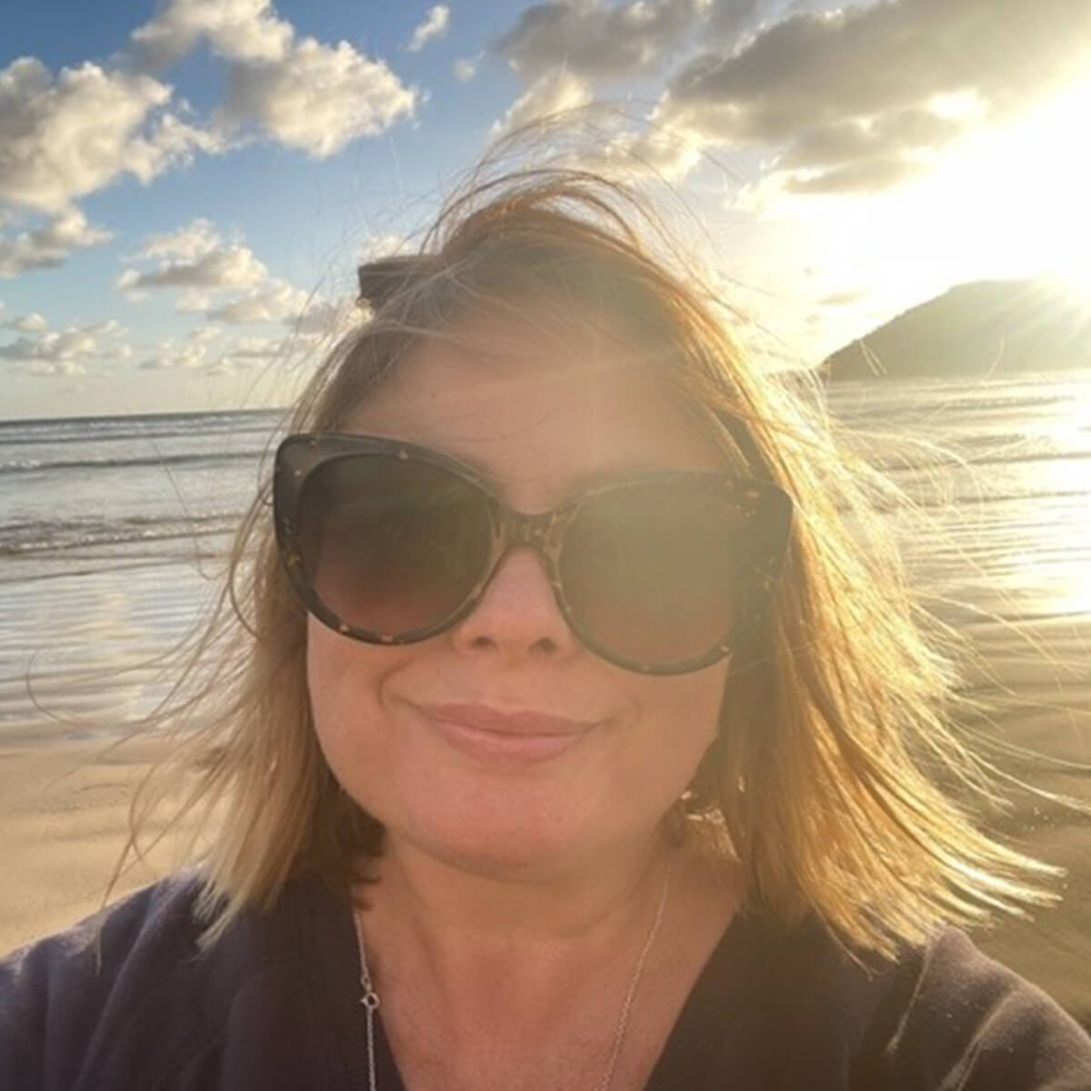 A woman with sunglasses stands on a beach, with windblown hair and a sunset in the background.