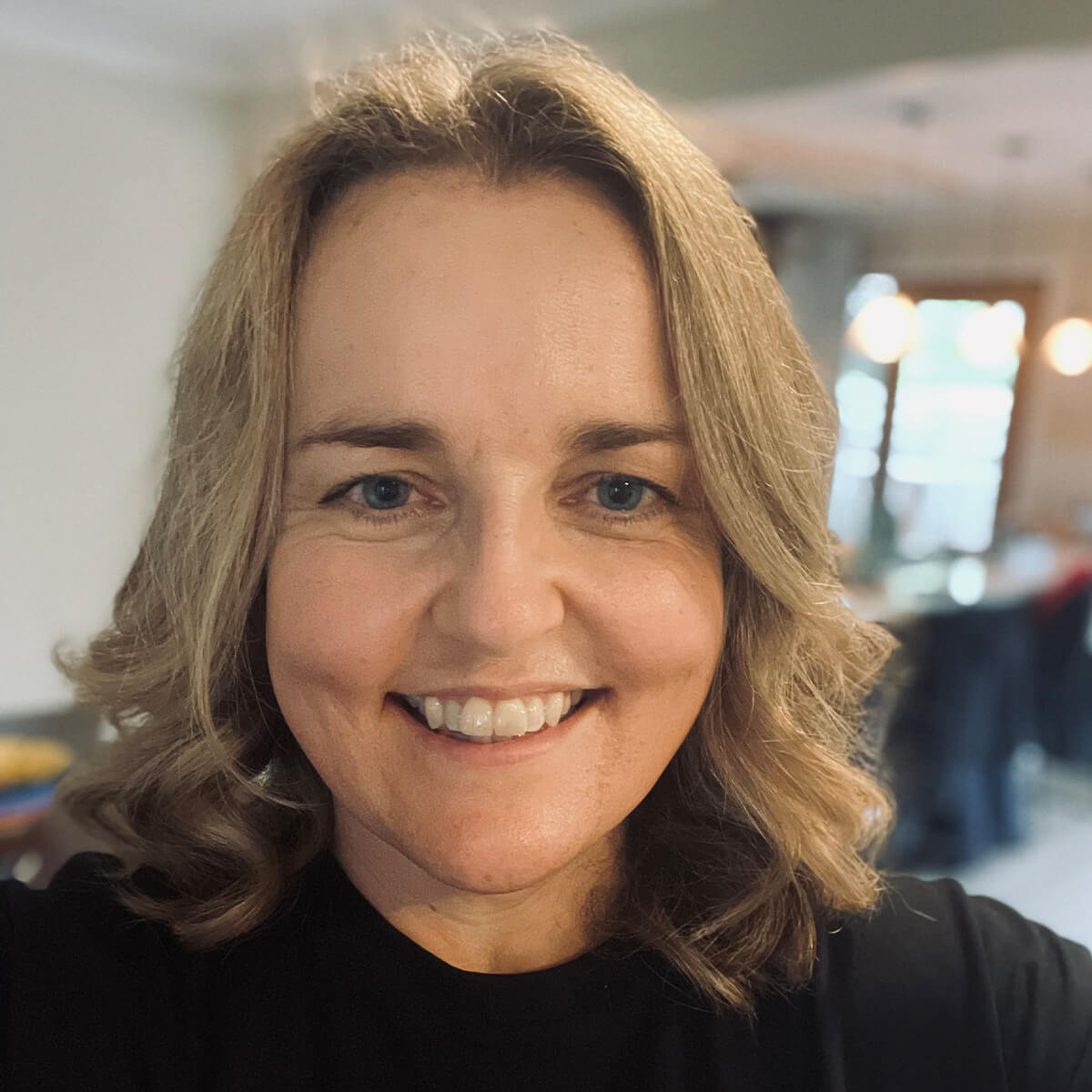A smiling woman with shoulder-length wavy hair and blue eyes, wearing a black shirt, in a well-lit indoor setting.