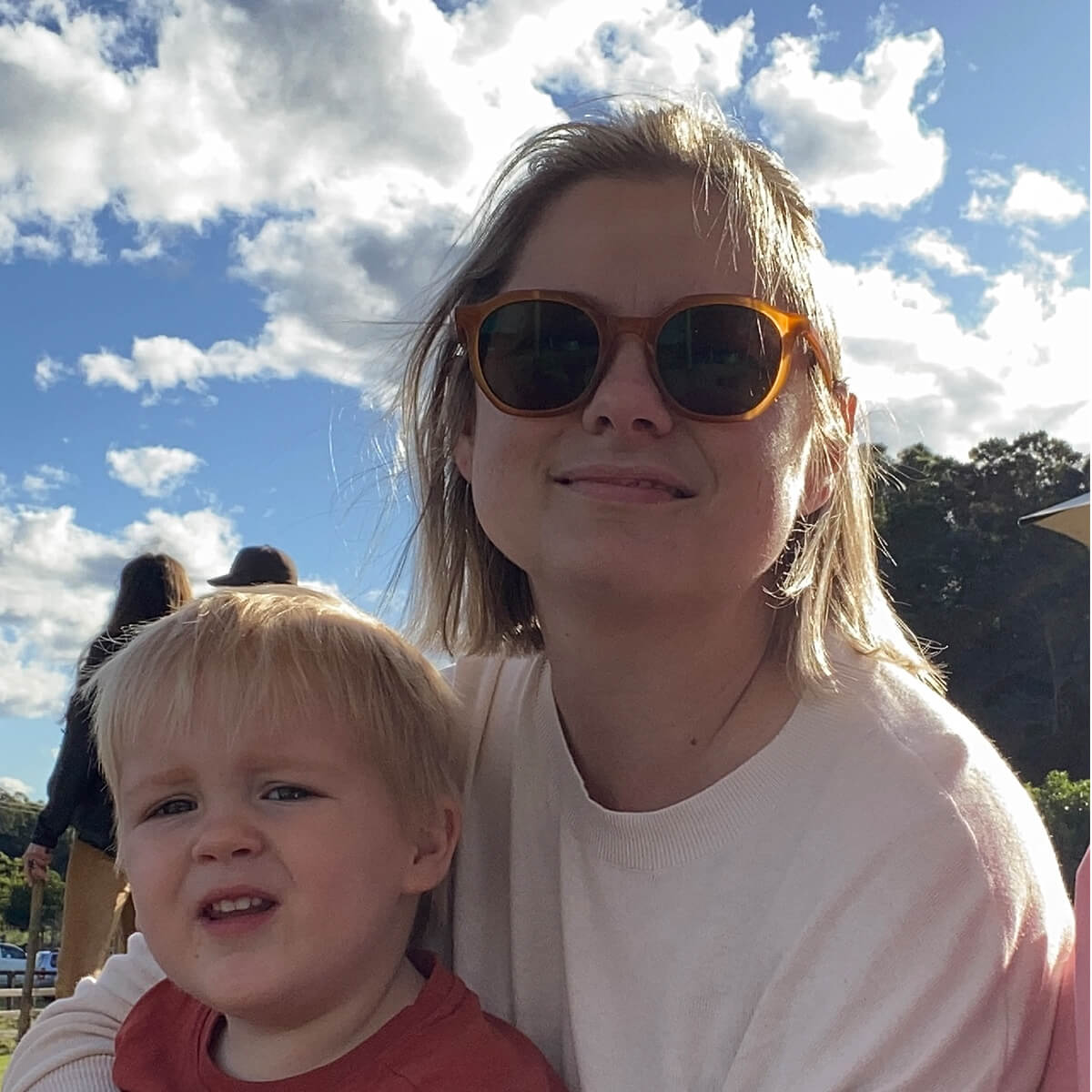A woman with sunglasses smiles while holding a young boy, both outdoors with a blue sky and clouds in the background.