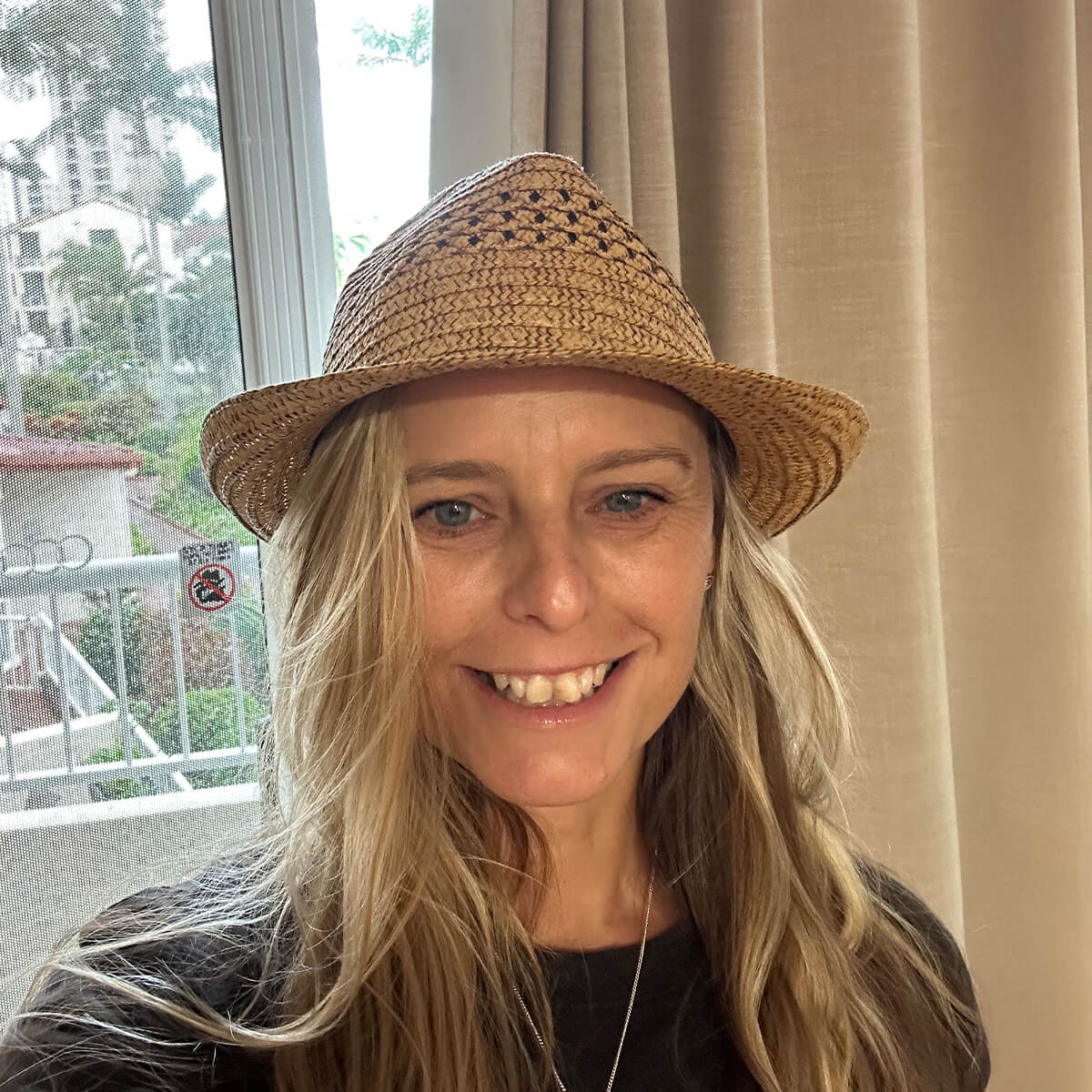 A woman with long hair smiles while wearing a straw hat, standing near a window with curtains.