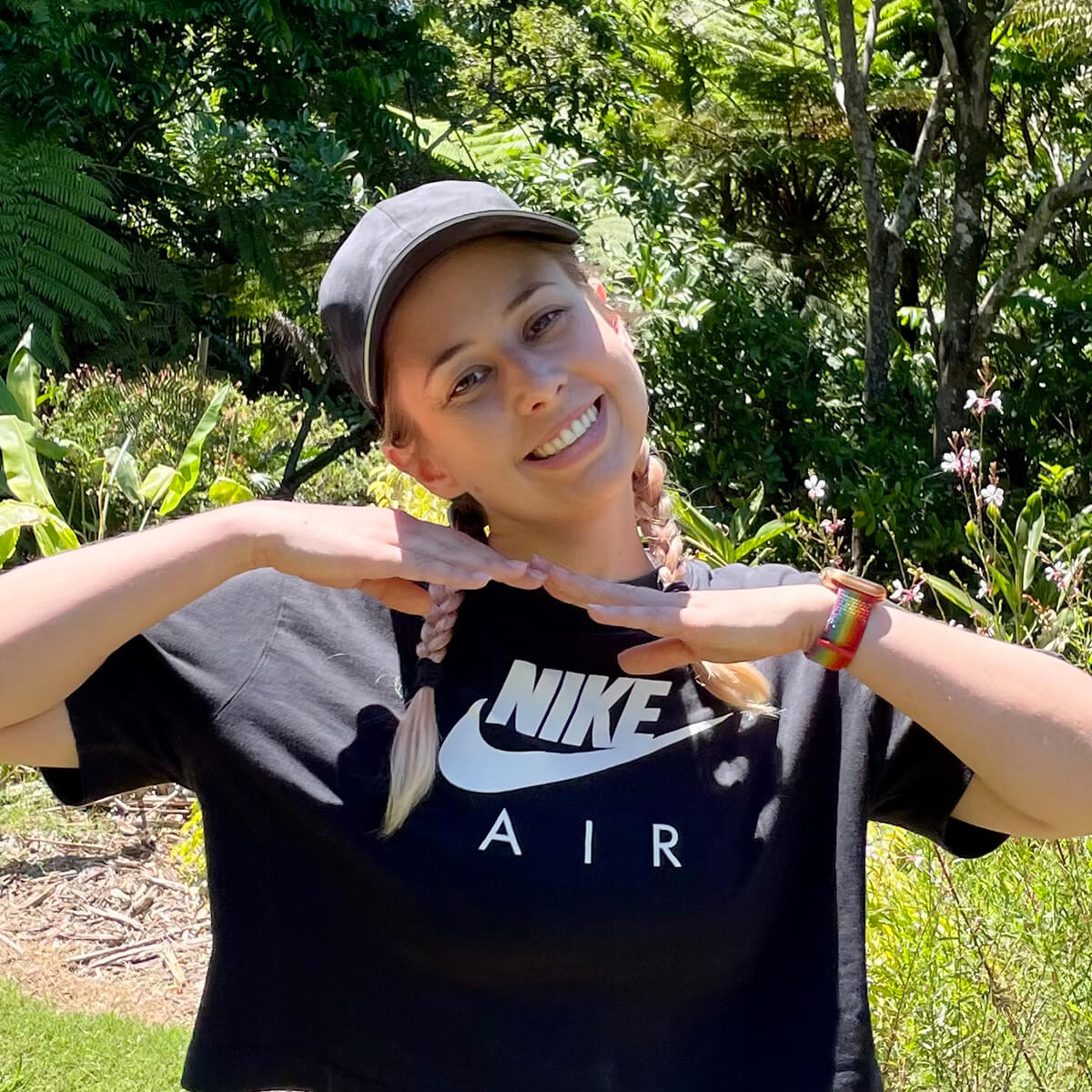 Smiling woman with braided hair in a black Nike shirt poses outdoors among greenery.