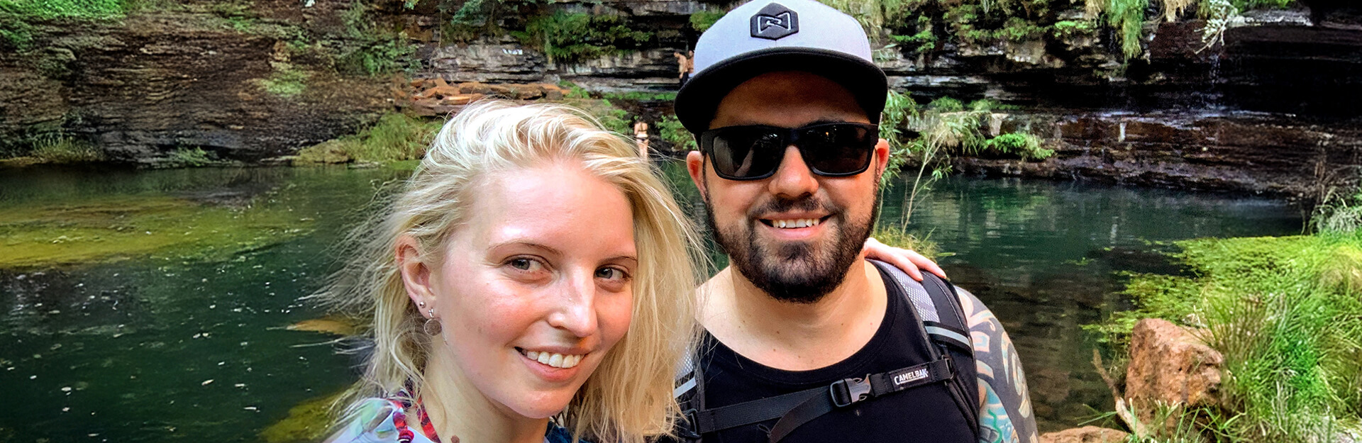 A couple smiles for a selfie by a serene green pond surrounded by rocky cliffs and lush vegetation.