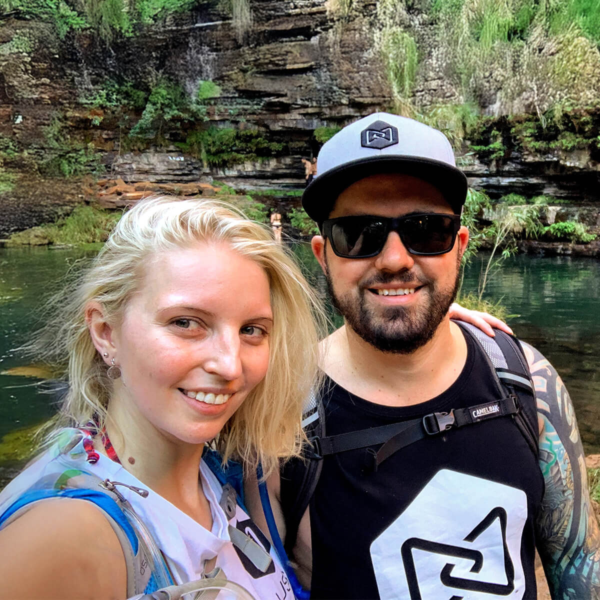 A man and a woman smile for a selfie by a serene water body surrounded by lush greenery and rocky cliffs.