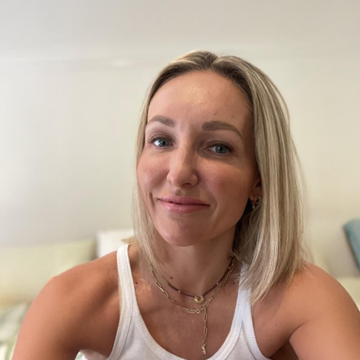 A woman with blonde hair and a white tank top smiles at the camera, sitting in a softly lit room.