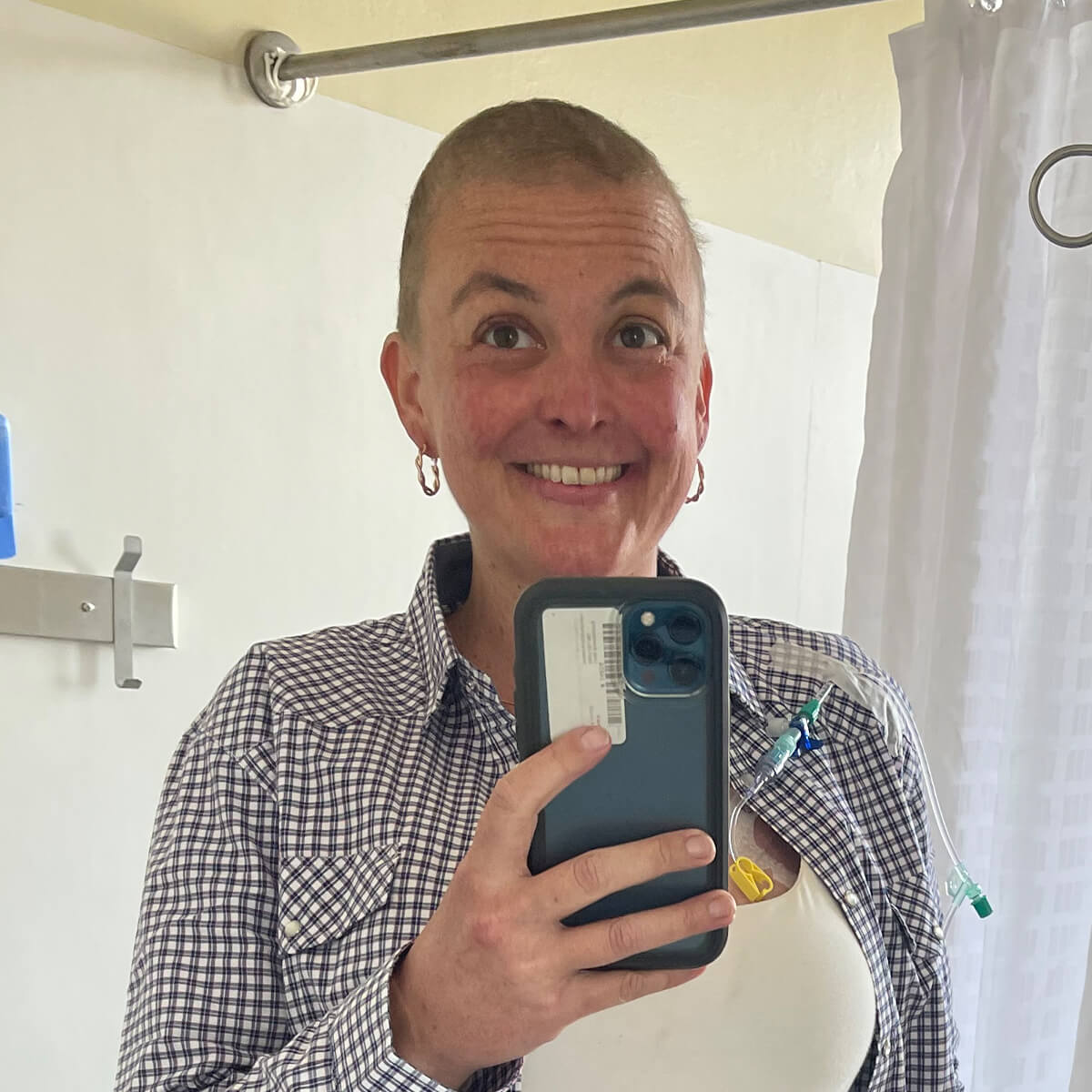 A person with a shaved head smiles in a mirror, wearing a checkered shirt and holding a phone. Medical equipment is visible.