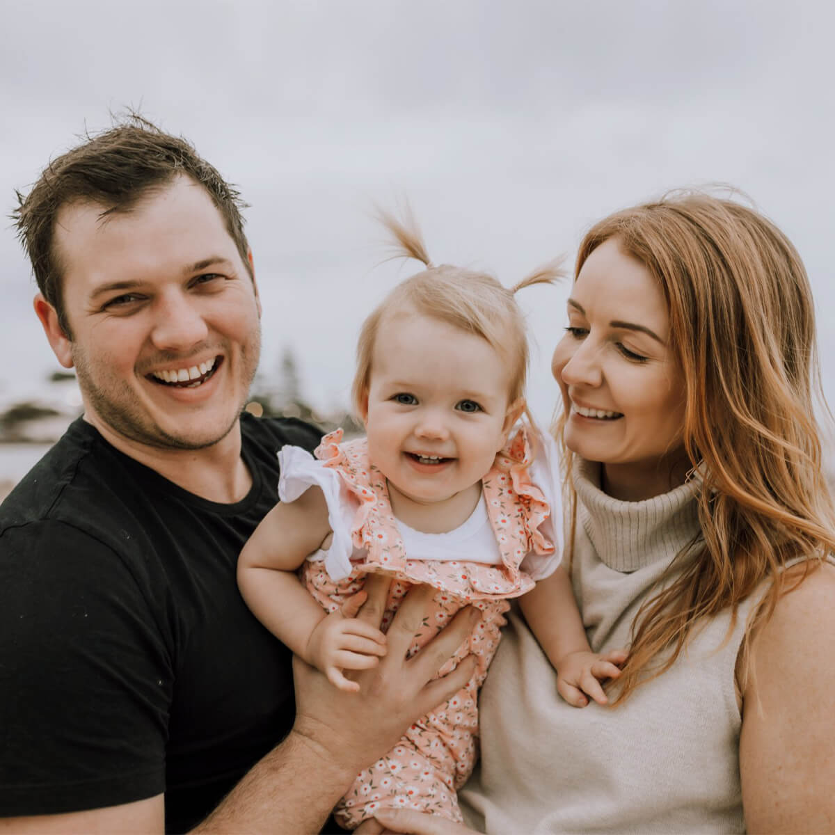 A smiling family of three, with a man holding a toddler girl, and a woman beside them, all outdoors.