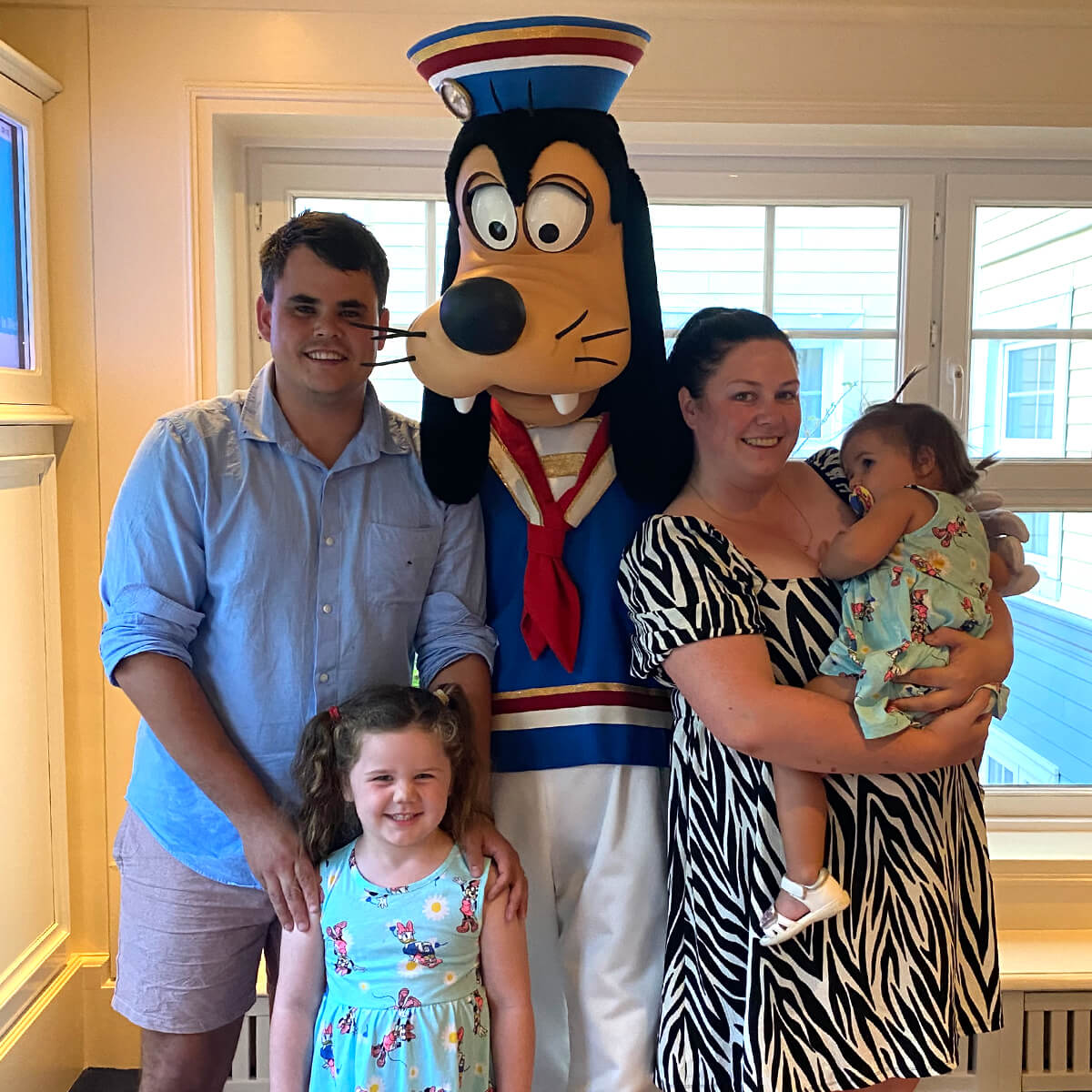 A family poses with a cartoon character in a colorful outfit, smiling in a well-lit indoor setting.