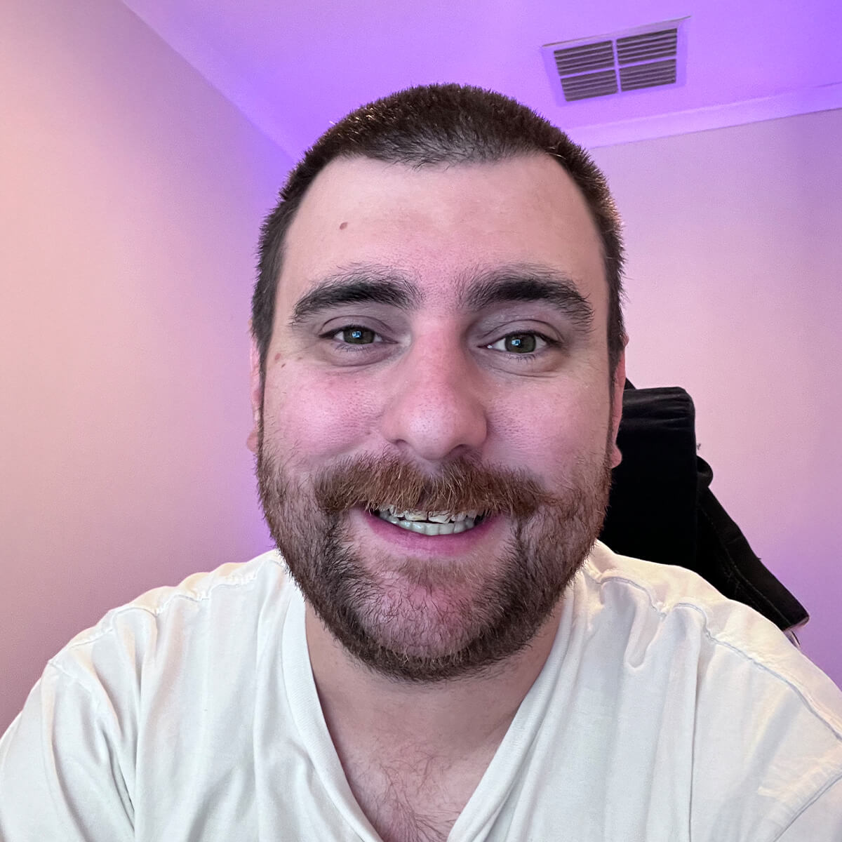 A smiling man with a beard and short hair is sitting in front of a light purple background.