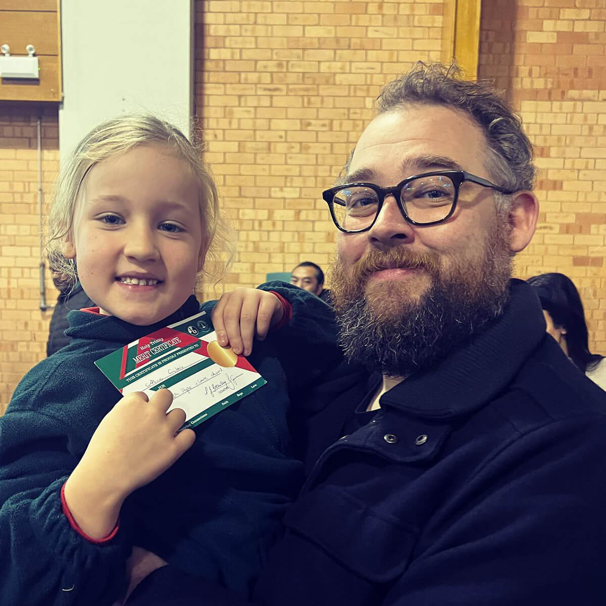 A smiling girl holds a certificate while posing with a bearded man in a crowd. They are indoors with a brick wall background.