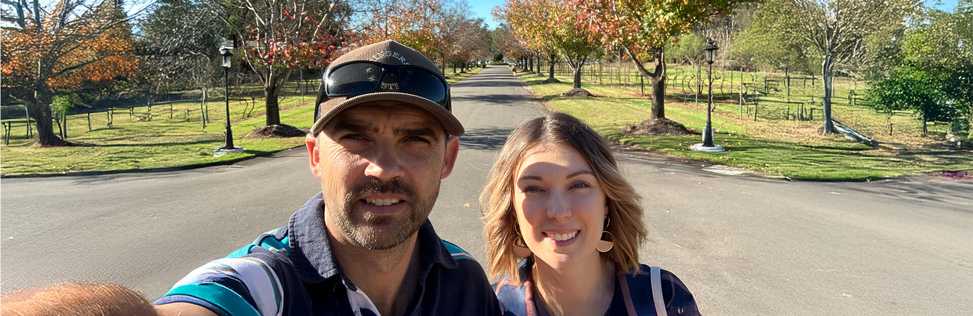 A man and woman take a selfie on a tree-lined road with autumn foliage and grassy areas.