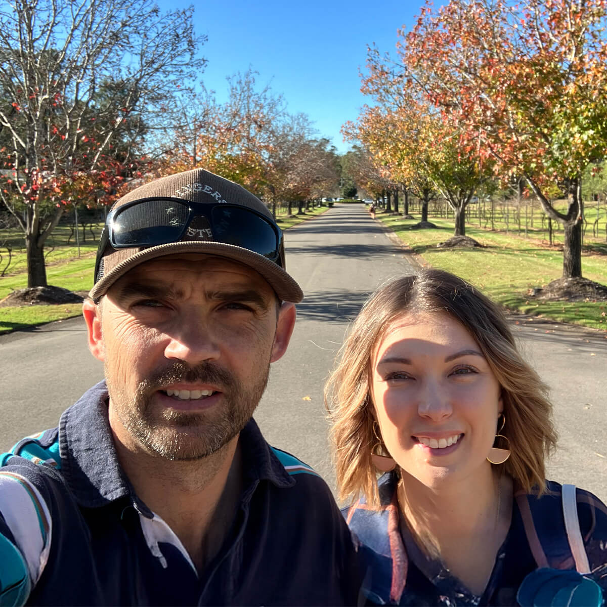 A man and a woman pose for a selfie on a tree-lined road with autumn foliage under a clear blue sky.