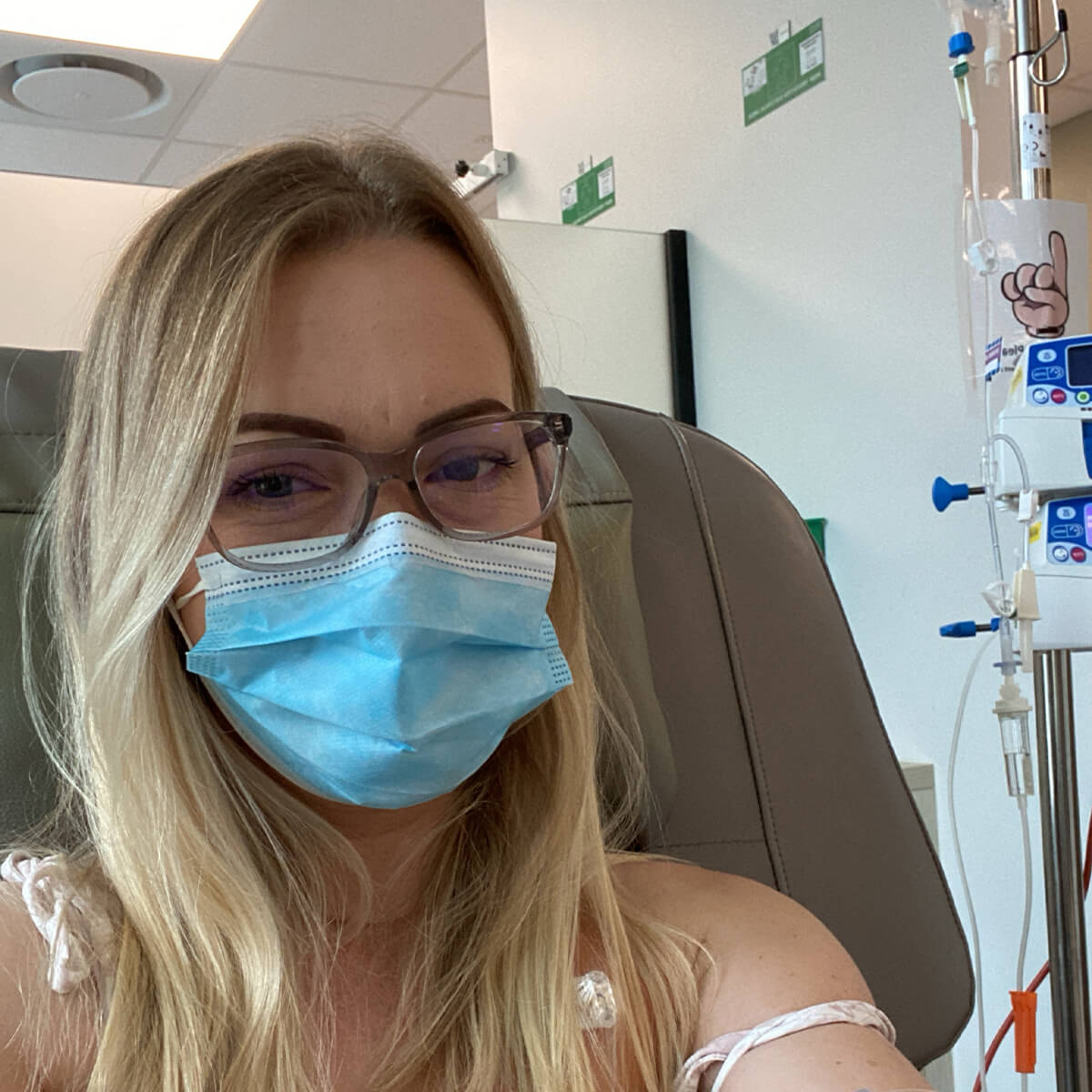 A woman with long blond hair and glasses wearing a blue mask sits in a medical setting next to an IV stand.