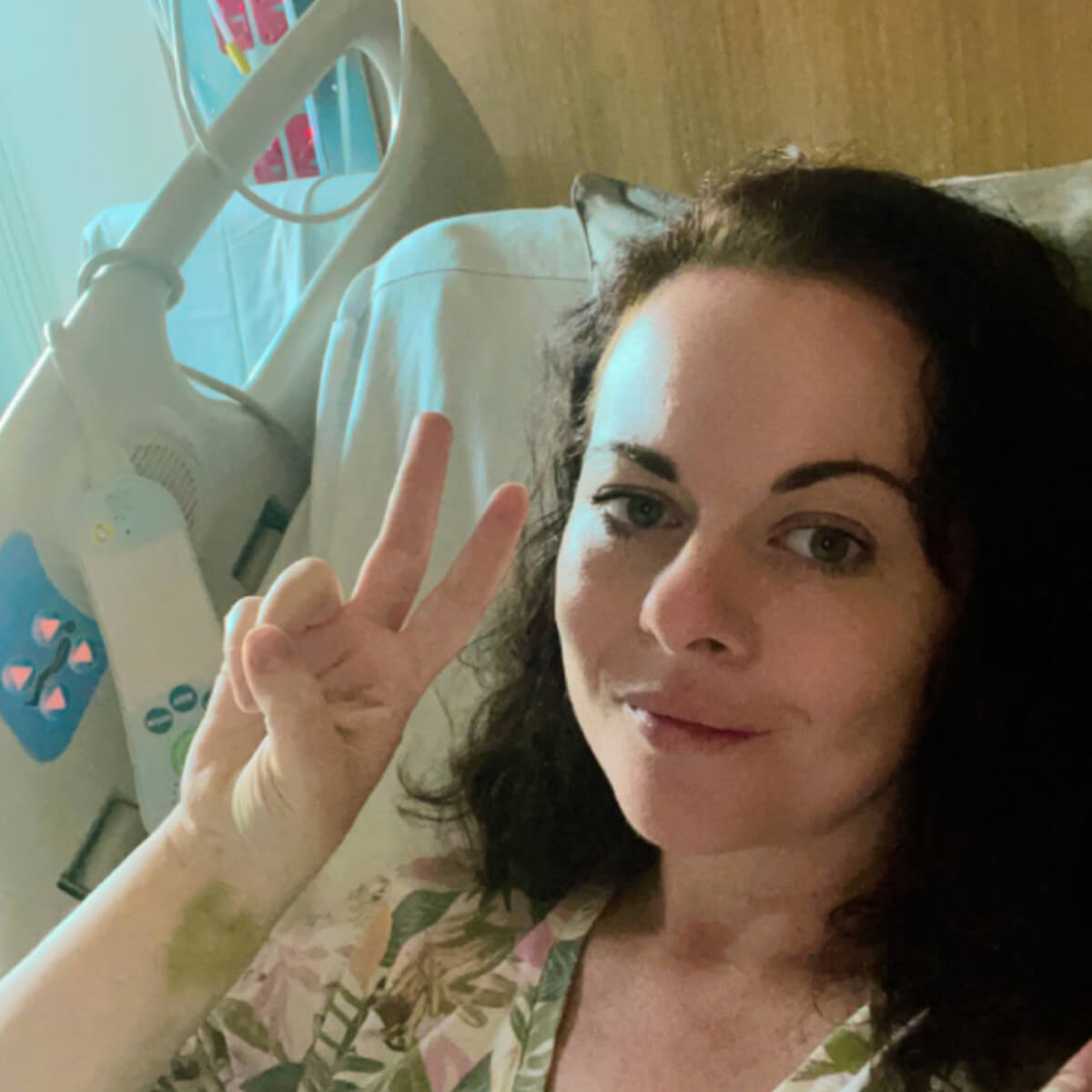 A person in a hospital bed smiles and makes a peace sign with their hand, wearing a floral-patterned hospital gown.